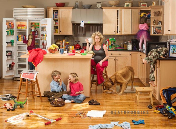 Уставшая беременная женщина сидит на кухне в страшном беспорядке. Мальчик и девочка едят торт на полу, мальчик лезет головой в микроволновку, девочка в шкаф, еще одна девочка в холодильник