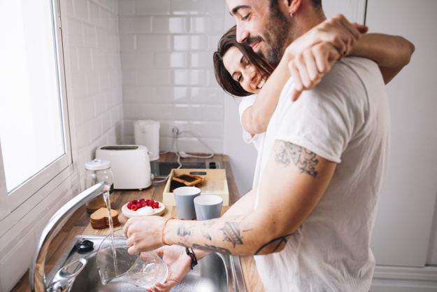 Мужчина моет посуду, его обнимает женщина, оба улыбаются